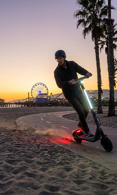 Person riding an electric scooter on a beach path at sunset with a Ferris wheel in the background.