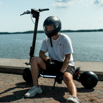 Person wearing a full-face helmet sitting on an electric scooter by the water.